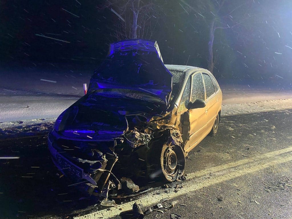 Rozbity samoch&oacute;d na drodze krajowej po wypadku, nocą, z uszkodzeniami na przednim i bocznym boku.