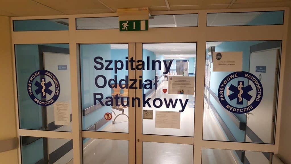Drzwi wejściowe do oddziału ratunkowego w szpitalu w Olsztynie, z napisem 'Szpitalny Oddział Ratunkowy' i symbolami medycznymi.