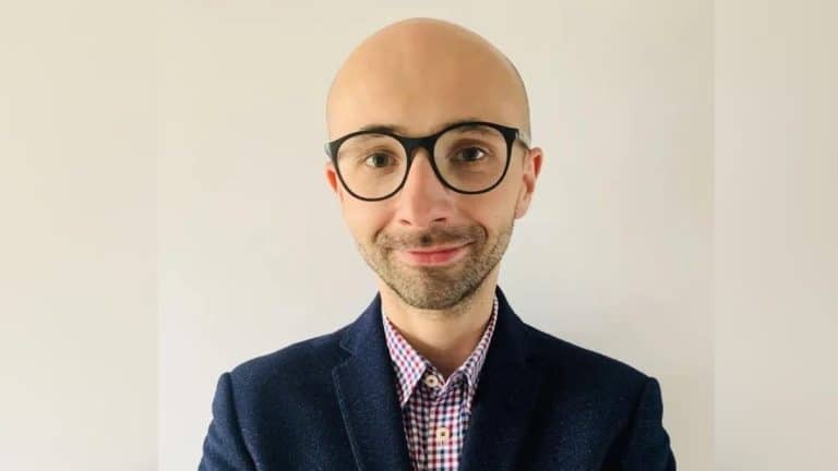 Mężczyzna w okularach, uśmiechnięty, w marynarce i koszuli, na jasnym tle.