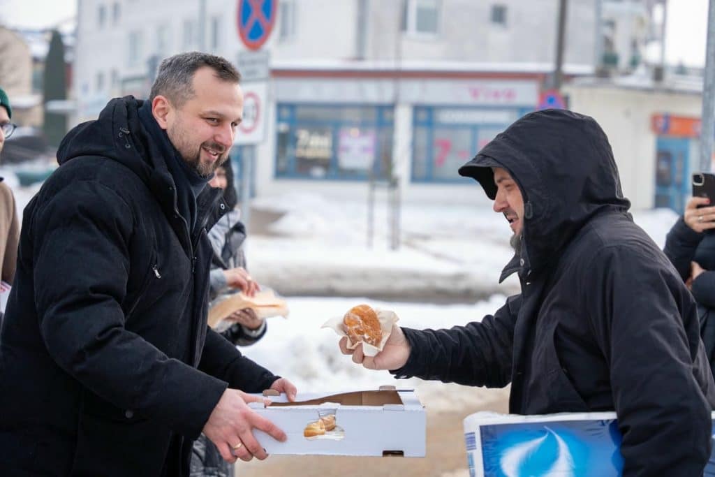 Prezydent Olsztyna rozdaje pączki na ulicy w Tłusty Czwartek, uśmiechając się do mieszkańc&oacute;w.