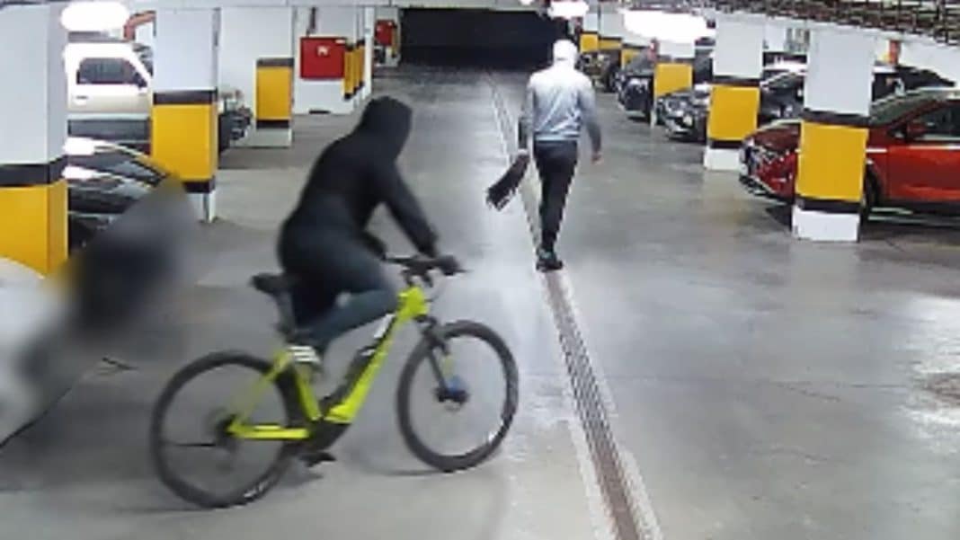 Nagranie z monitoringu pokazujące dw&oacute;ch mężczyzn w kapturach, jeden na rowerze, drugi idący z torbą, w parkingu.