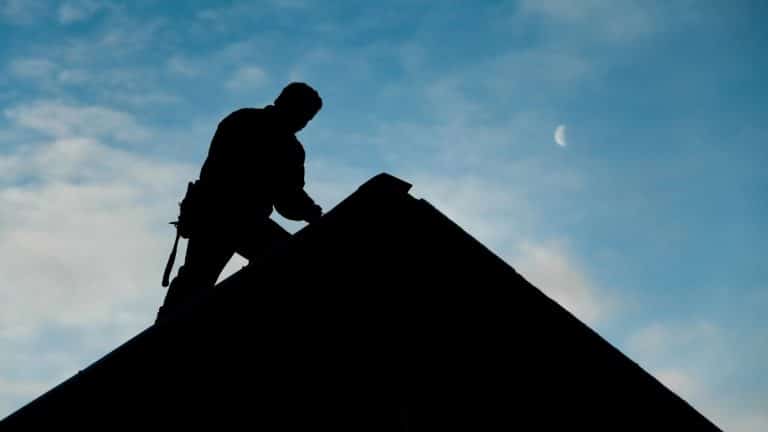 Człowiek montujący pokrycie dachowe na stromym dachu w trakcie pracy na zewnątrz, w tle niebo z księżycem.