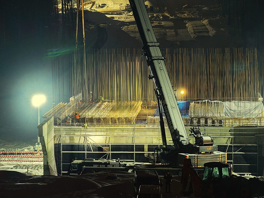 Prace budowlane na placu budowy w nocy, widoczny ciężki sprzęt i konstrukcje stalowe.