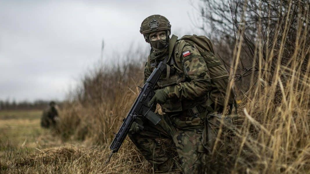 Żołnierz w maskowaniu, ukryty w trawie, z bronią, na tle jesiennego krajobrazu.