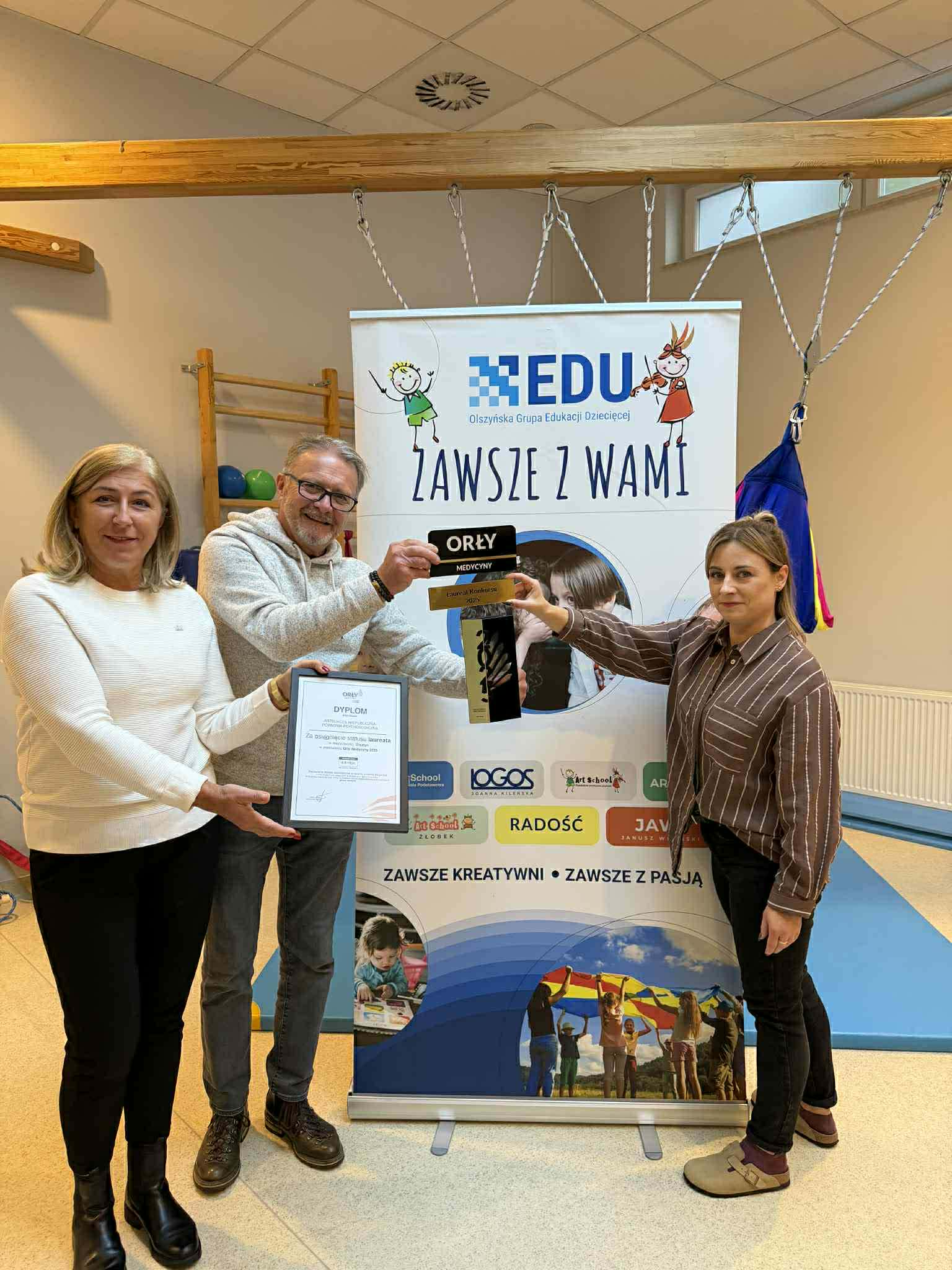 Trzy osoby trzymają nagrody i certyfikat na tle plakatu promującego dni otwarte przedszkola i żłobka w Olsztynie.