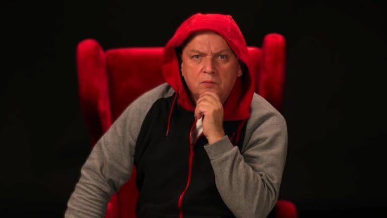 Mężczyzna w czerwonej bluzie z kapturem siedzi na czerwonym fotelu, patrząc poważnie.