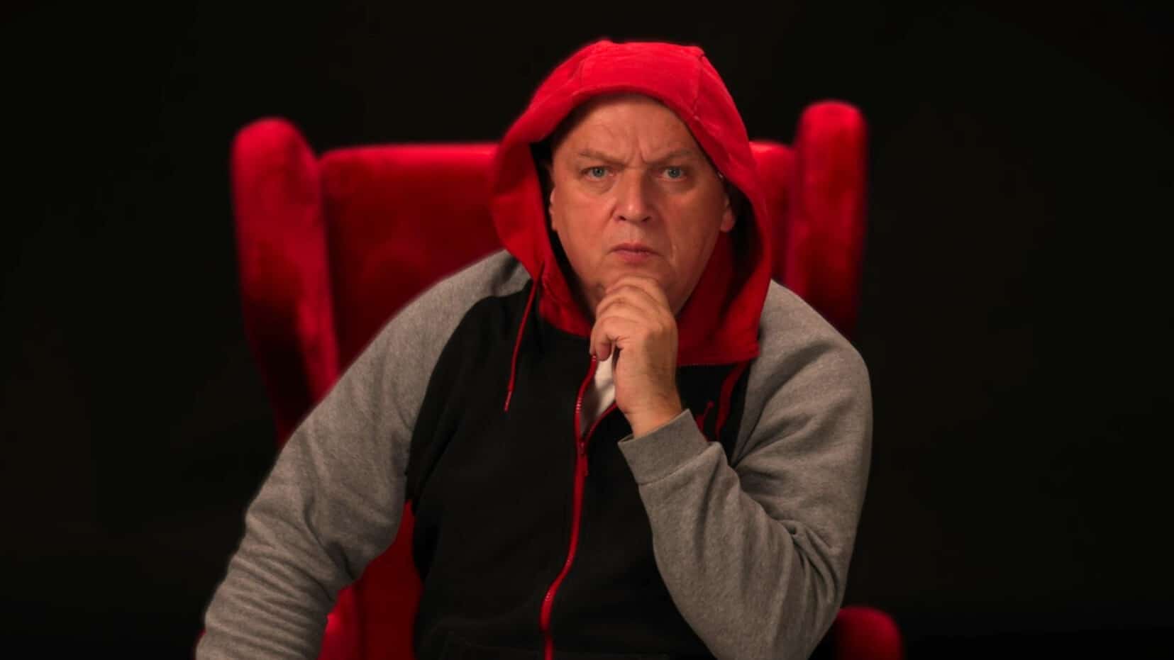Mężczyzna w czerwonej bluzie z kapturem siedzi na czerwonym fotelu, patrząc poważnie.