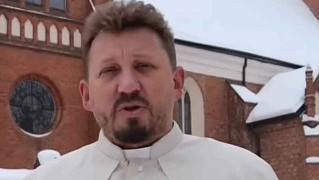 Ksiądz przemawia przed zabytkową bazyliką w Gietrzwałdzie, w tle widoczne ściany i dach kościoła.