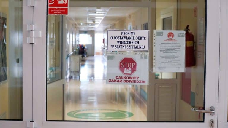 Szpital z zamkniętymi drzwiami, na kt&oacute;rych wiszą oznaczenia o zakazie odwiedzin i konieczności zakrywania twarzy.