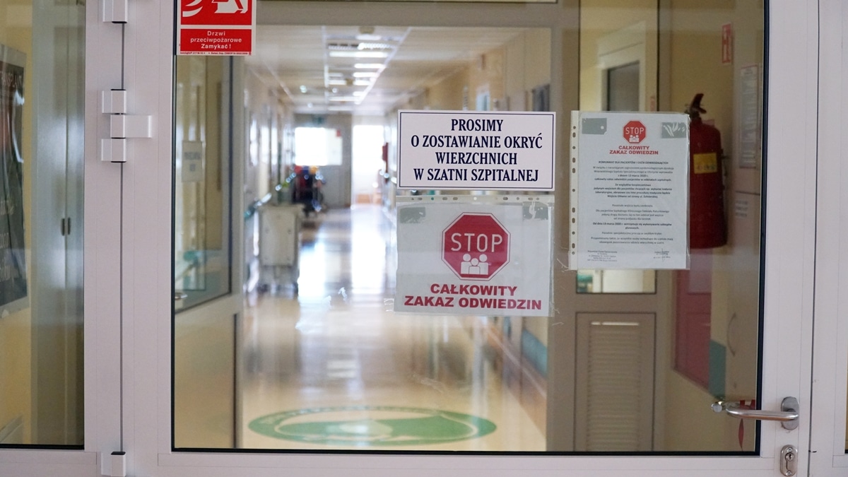 Szpital z zamkniętymi drzwiami, na których wiszą oznaczenia o zakazie odwiedzin i konieczności zakrywania twarzy.