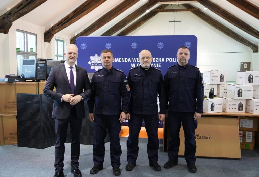 Policjanci i przedstawiciel władz na tle tablicy informacyjnej, obok sprzęt i opakowania.