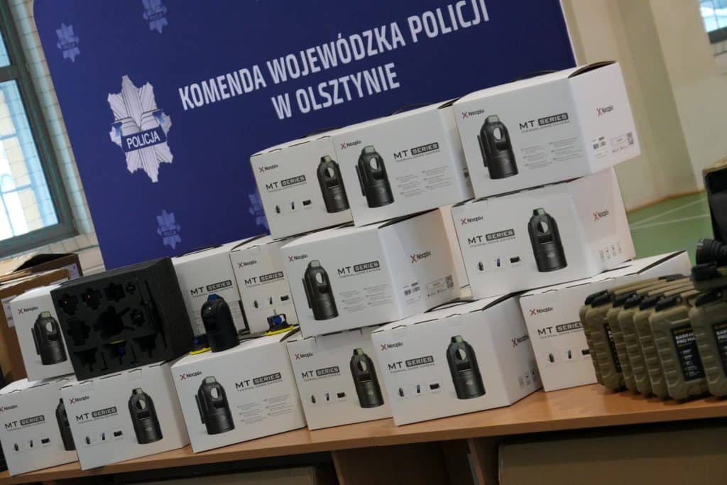 Nowoczesny sprzęt policyjny na stole, w tle plakat z napisem 'Komenda Wojew&oacute;dzka Policji w Olsztynie'.