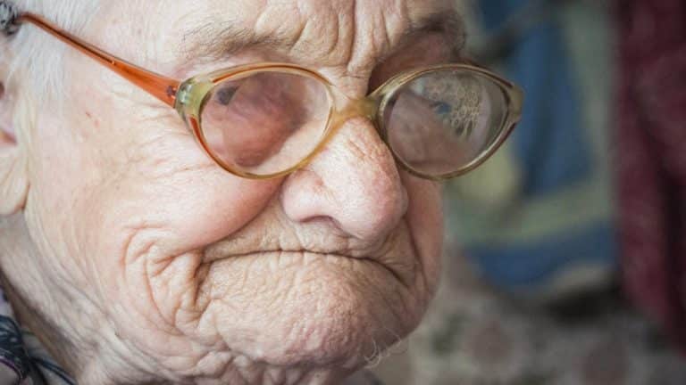Zbliżenie na twarz starszej kobiety z okularami, widoczne zmarszczki i delikatny uśmiech.