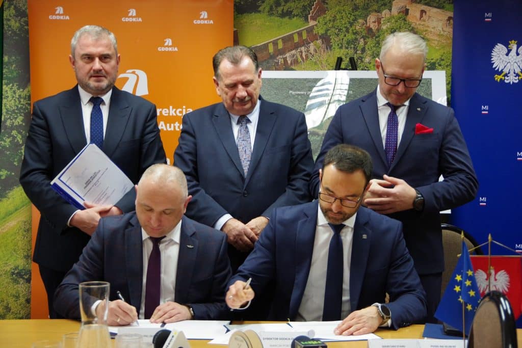 Podpisanie umowy na budowę wiaduktu drogowego w Iławie, grupa mężczyzn w garniturach podczas ceremonii.