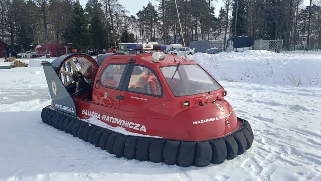 Pojazd ratowniczy na zamarzniętym jeziorze w zimowej scenerii, patrol policyjny w tle.