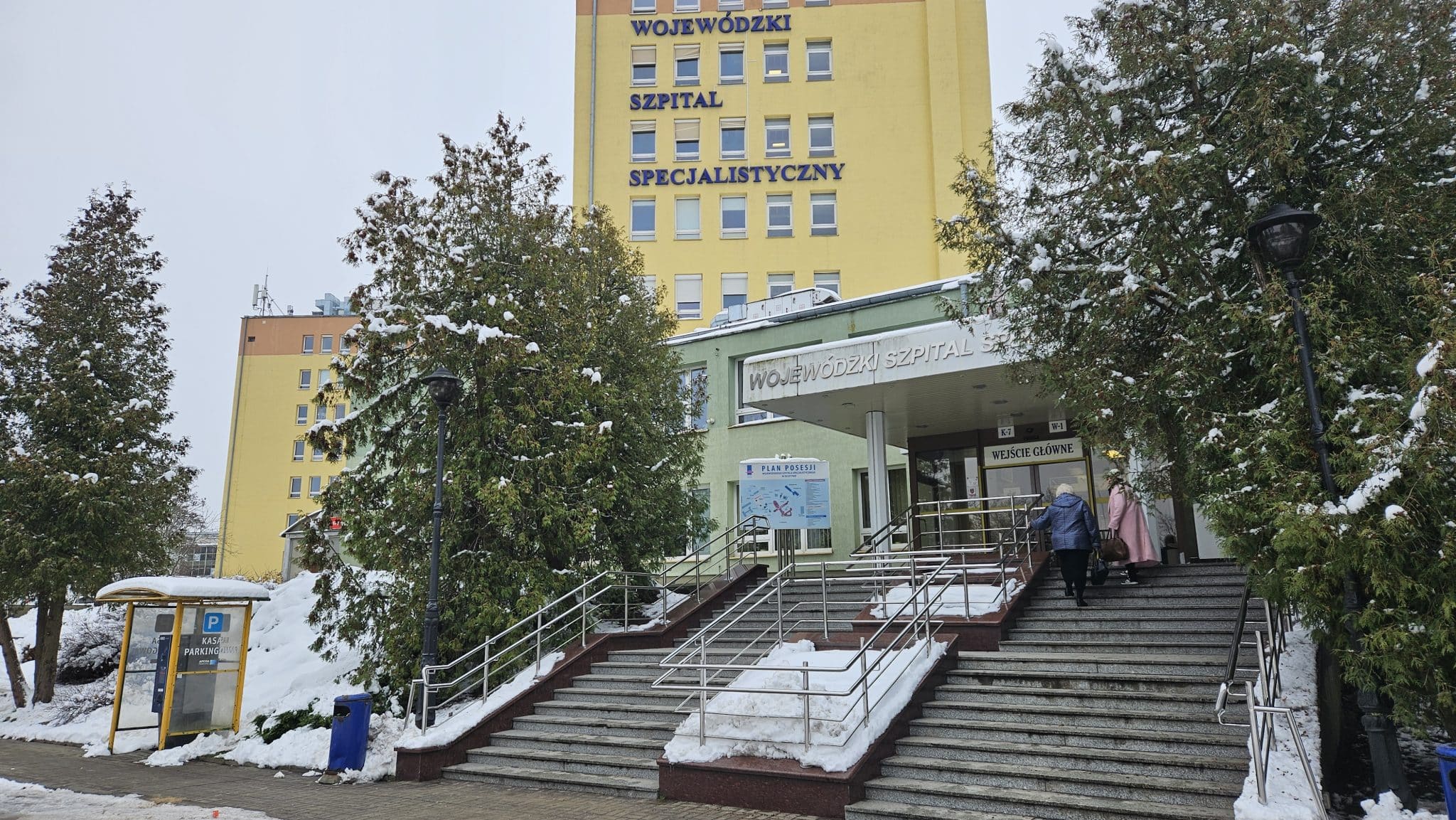 Widok wejścia do Wojewódzkiego Szpitala Specjalistycznego w Olsztynie zimą, z zaśnieżonymi drzewami i schodami.
