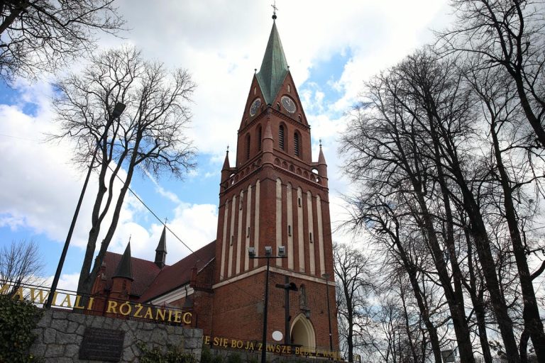 Widok zabytkowej bazyliki w Gietrzwałdzie z pękającymi ścianami i drzewami wokół, pod chmurami.