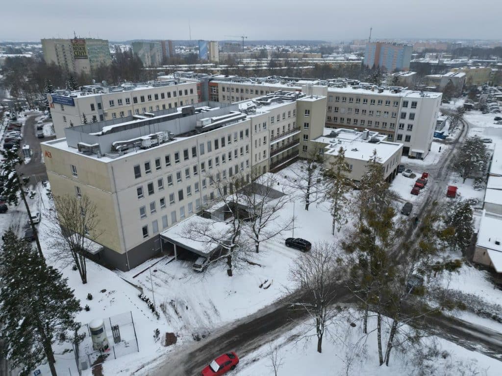 Widok na szpital dziecięcy w Olsztynie pokryty śniegiem, budynki i drzewa w zimowej scenerii.