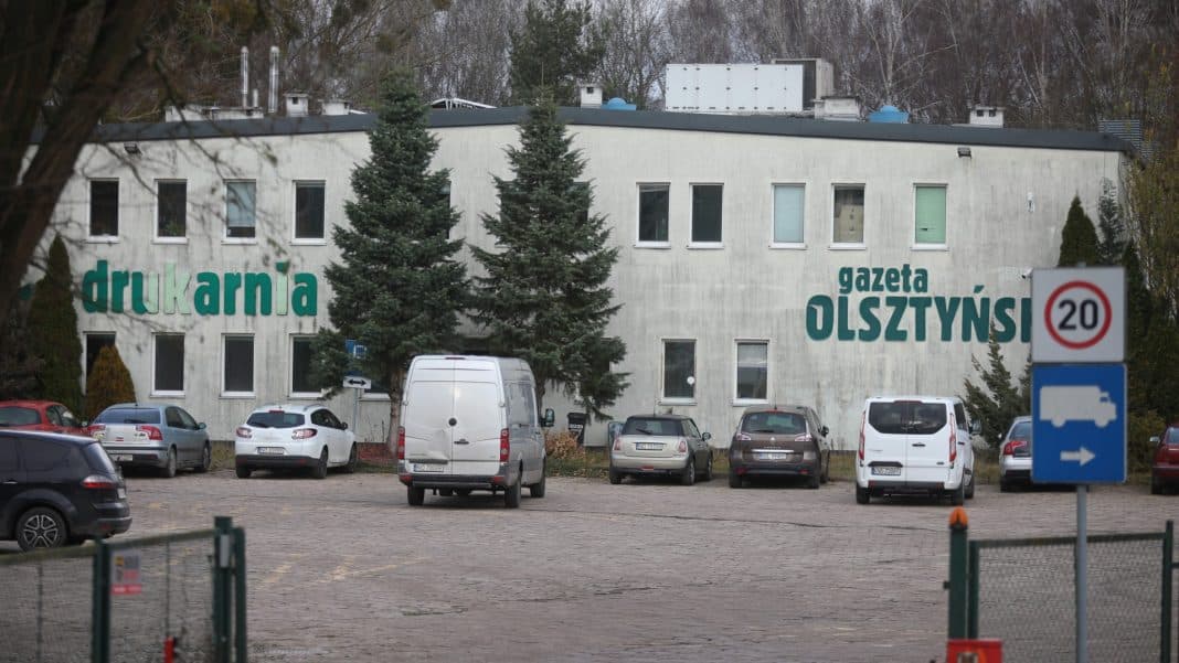 Widok na drukarnię i parking z samochodami, na budynku widnieje napis 'drukarnia gazeta Olsztyńska'.