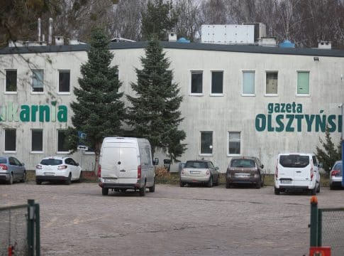 Widok na drukarnię i parking z samochodami, na budynku widnieje napis 'drukarnia gazeta Olsztyńska'.