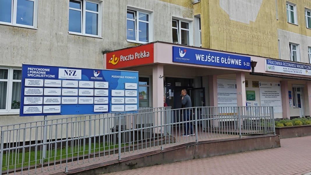 Widok wejścia do szpitala powiatowego z dużym niebieskim i czerwonym oznakowaniem.