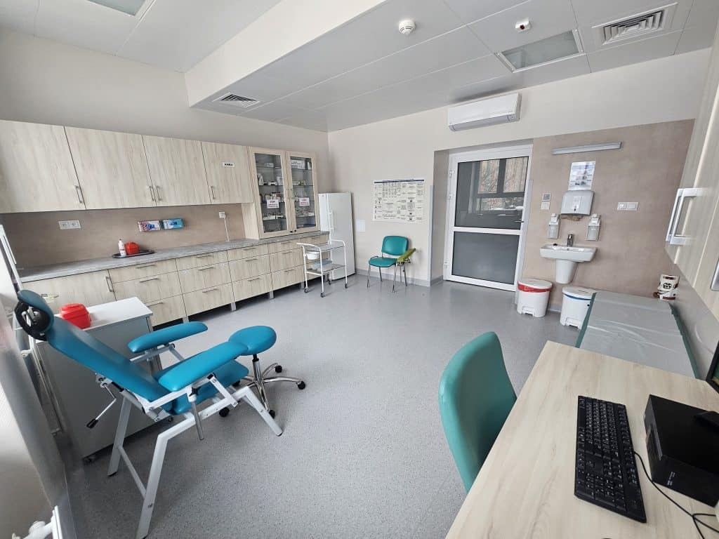 Nowoczesna sala szpitalna z ł&oacute;żkiem, biurkiem i sprzętem medycznym, jasne wnętrze, okno i sprzęt sanitarny.