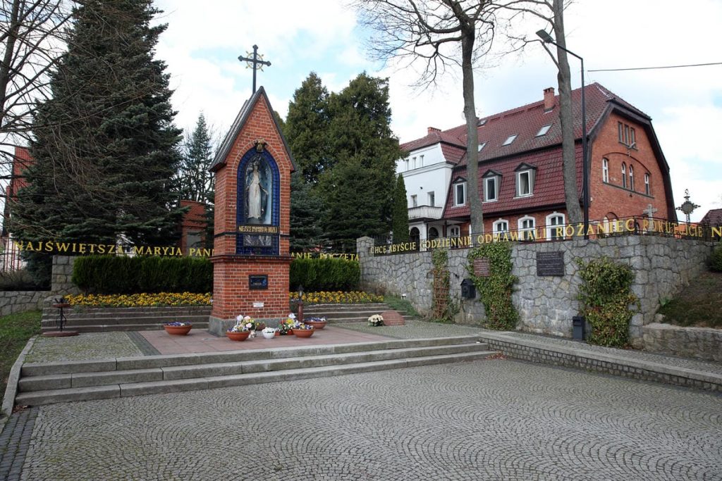 Zabytkowa bazylika w Gietrzwałdzie z pękającymi ścianami i kwiatami przed pomnikiem Matki Boskiej.