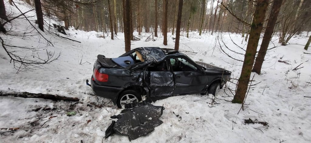 Uszkodzone czarne auto w lesie po wypadku, zniszczone drzwi i rozbity dach, wok&oacute;ł śnieg i drzewa.