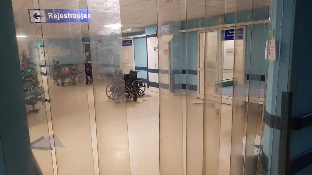 Widok na zamknięte drzwi wejściowe do szpitala z widocznymi w tle w&oacute;zkami inwalidzkimi i rejestracją.