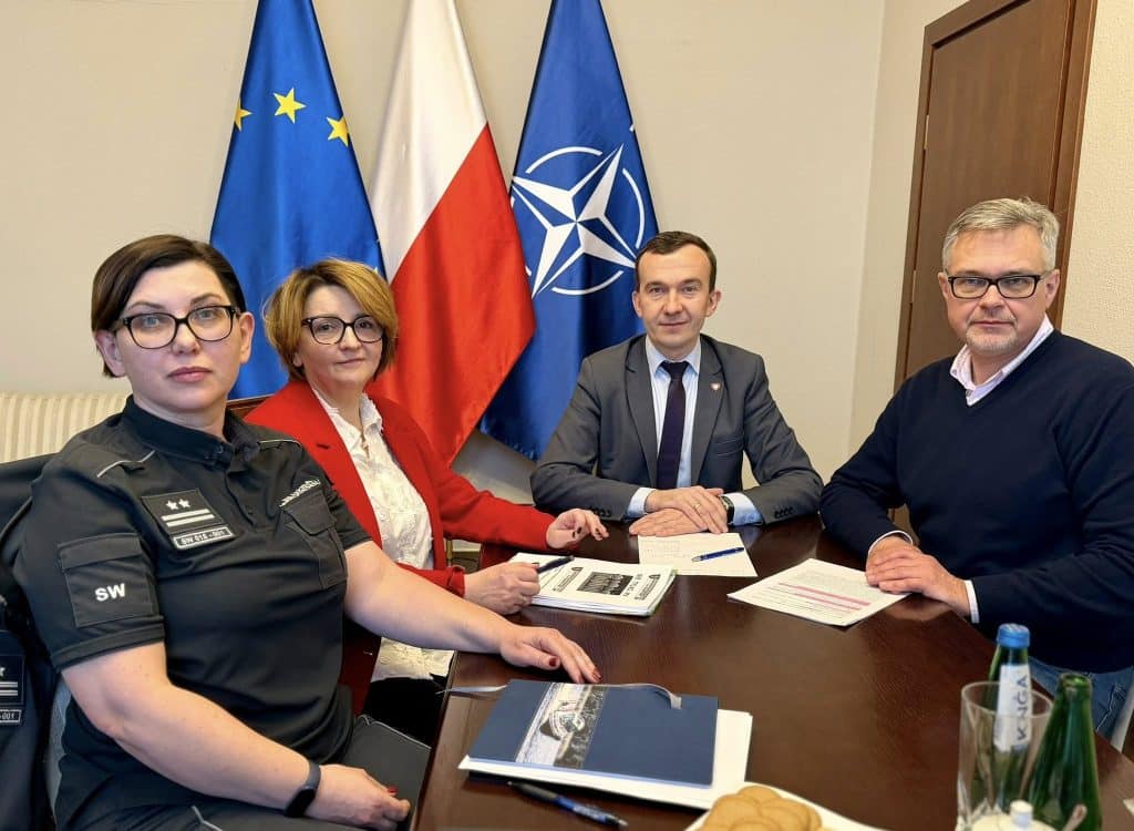 Grupa os&oacute;b podczas spotkania w urzędzie, z flagami UE, Polski i NATO w tle.