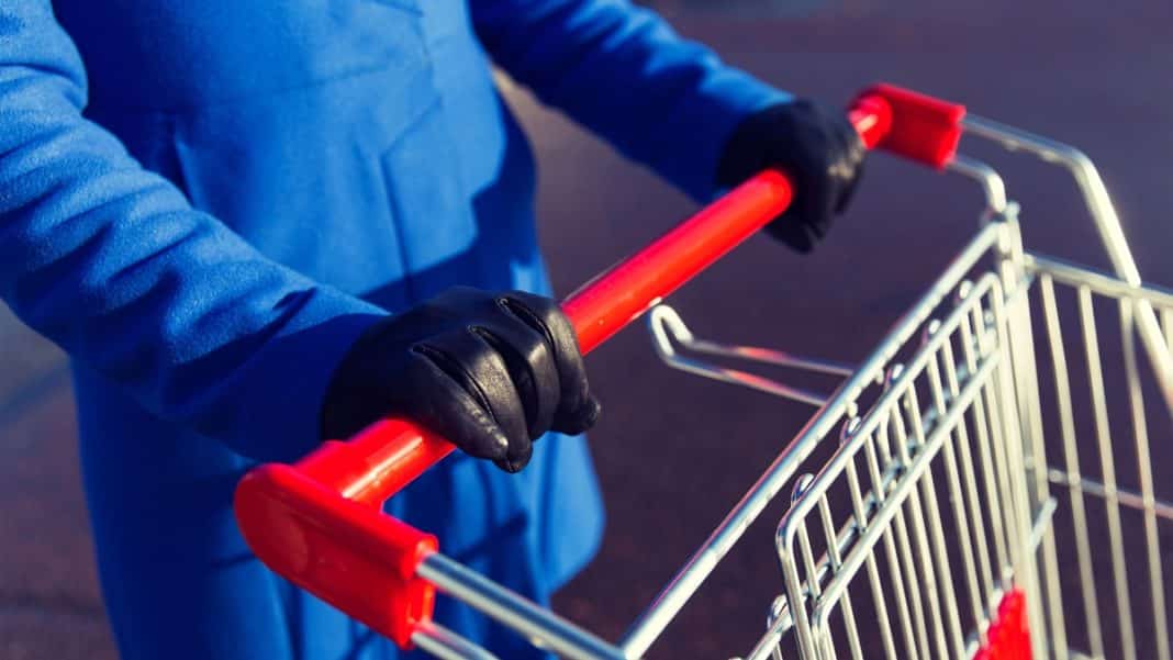 Osoba w niebieskim płaszczu i czarnych rękawiczkach trzymająca w&oacute;zek na zewnątrz sklepu, zbliżenie na ręce i w&oacute;zek.