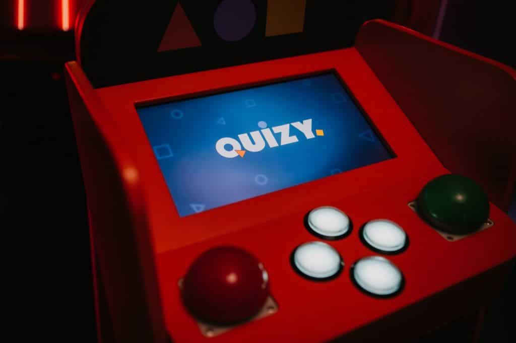 Automat do gry w quizy na tle ciemnego wnętrza, kolor czerwony i ekran z napisem 'Quizy'.