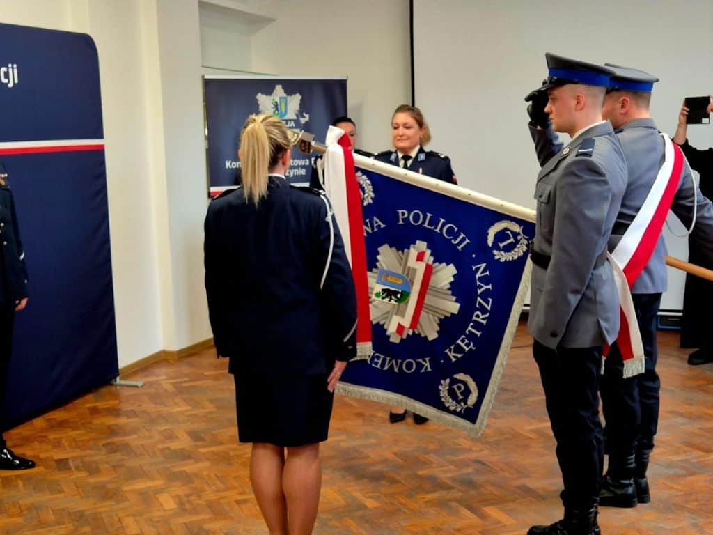 Policjanci podczas uroczystości z flagą, w tle plakaty i sala konferencyjna.