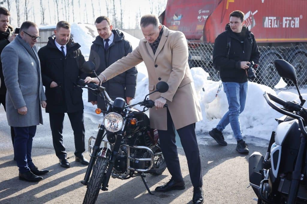 Mężczyzna w eleganckim płaszczu odkręca kierownicę motocykla podczas oficjalnego otwarcia filii WORD w Ełku, wok&oacute;ł grupa ludzi.