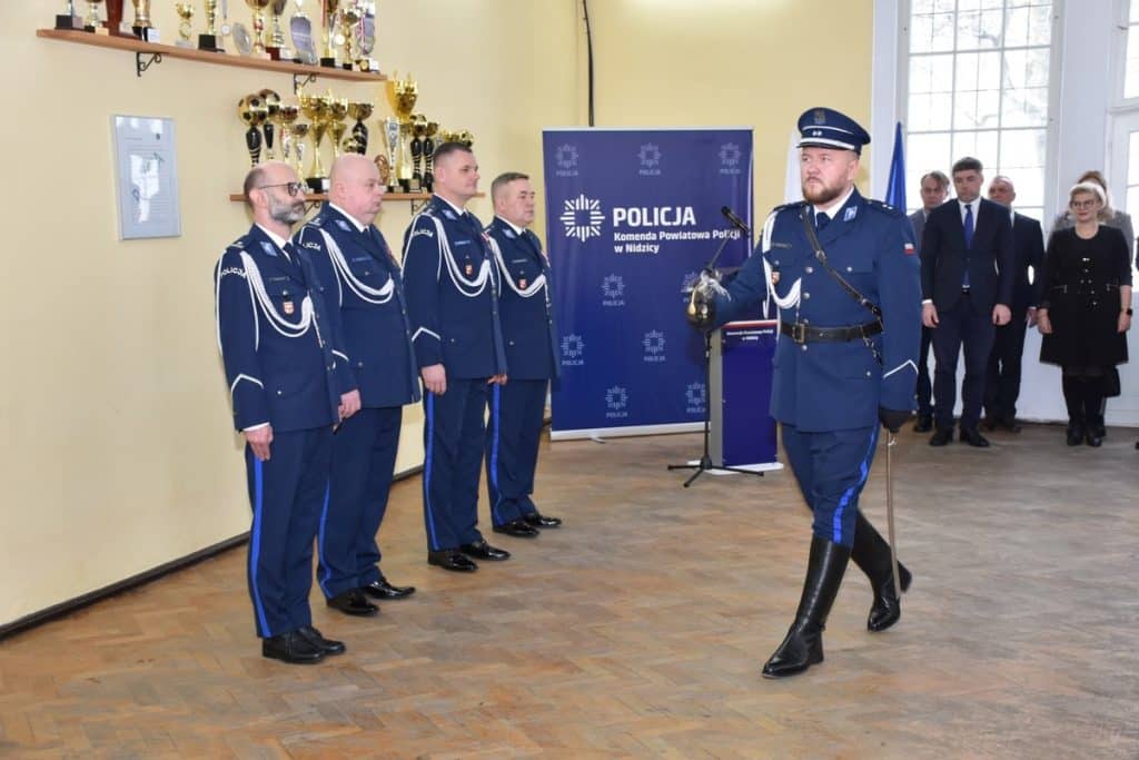 Policjanci w uniformach podczas uroczystości w komendzie w Nidzicy, na tle ściany z trofeami.