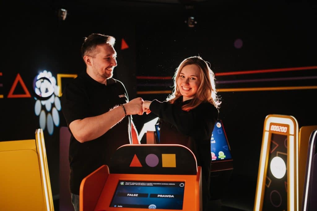 Dwoje ludzi uśmiecha się i trzyma za ręce w nowoczesnym lokalu rozrywkowym, w tle kolorowe światła i grafiki.