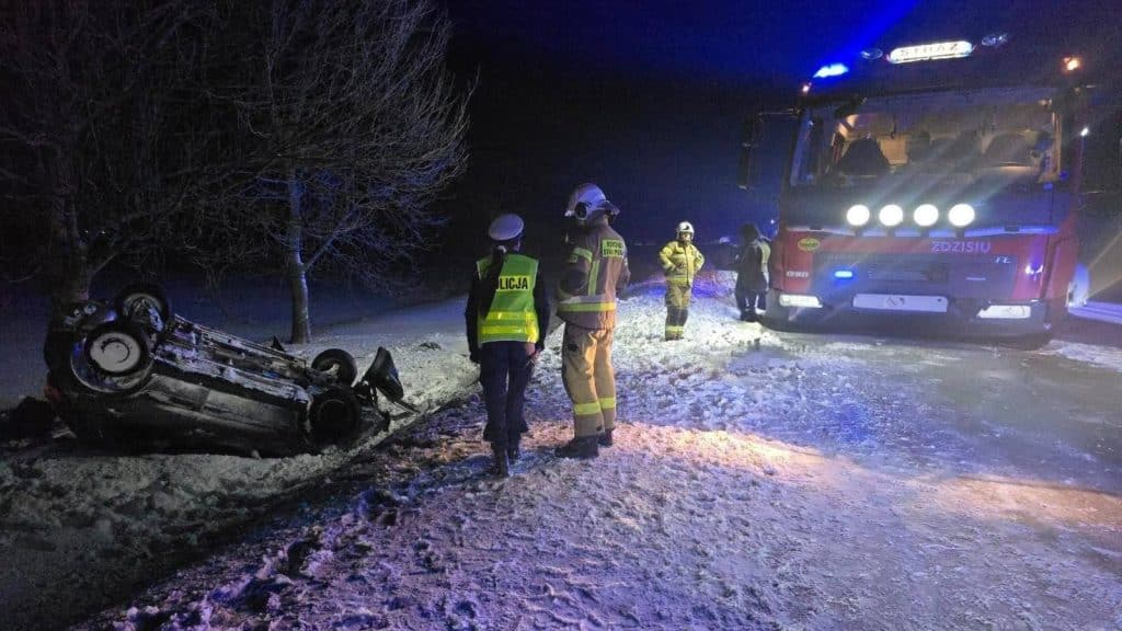 Samoch&oacute;d dachował na śnieżnej drodze, wok&oacute;ł strażacy i policjanci, nocne warunki, służby na miejscu.