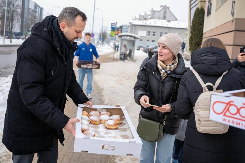 Prezydent Olsztyna rozdaje pączki na ulicy, rozmawia z mieszkańcami, w tle śnieg i przystanek autobusowy.