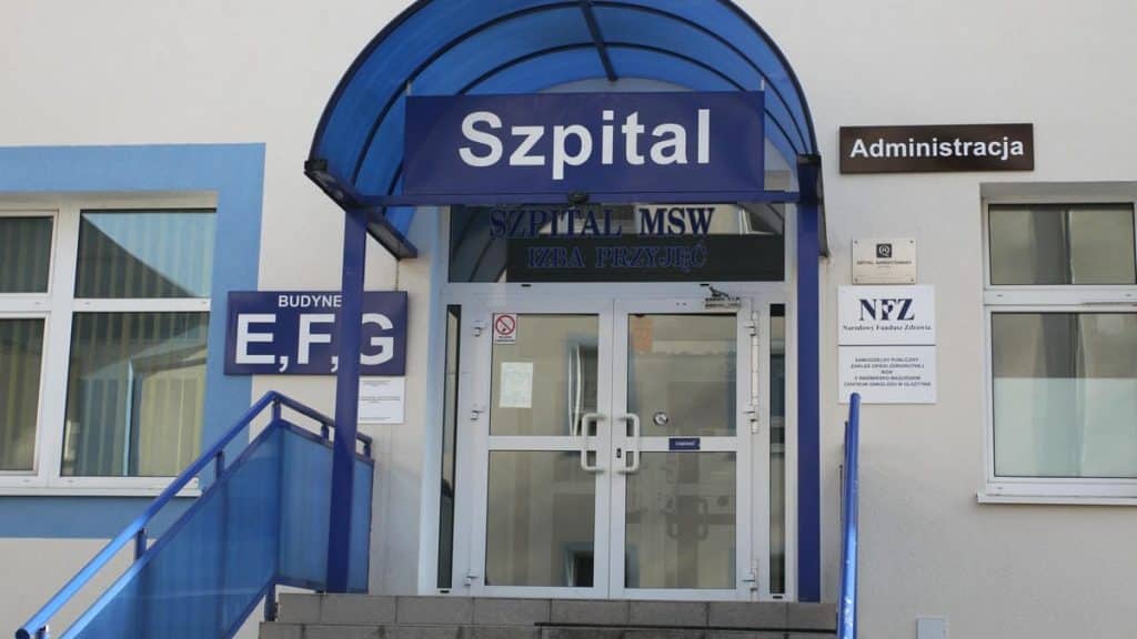 Wejście do szpitala z oznaczeniem 'Szpital' i administracją, widoczne drzwi wejściowe.