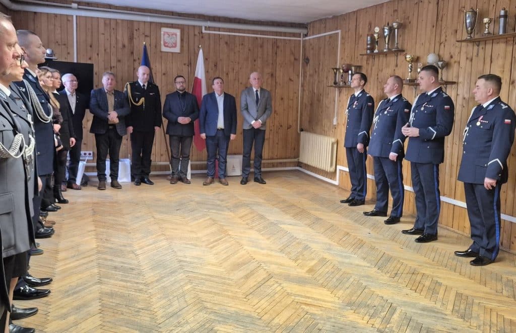 Policjanci i urzędnicy podczas oficjalnego spotkania w drewnianym pomieszczeniu, na ścianach trofea i flagi.