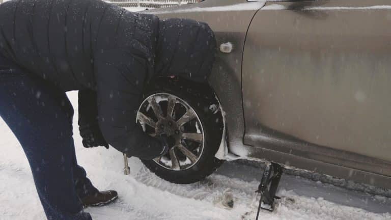 Mężczyzna wymieniający przebitą oponę w samochodzie na zaśnieżonej ulicy zimą.