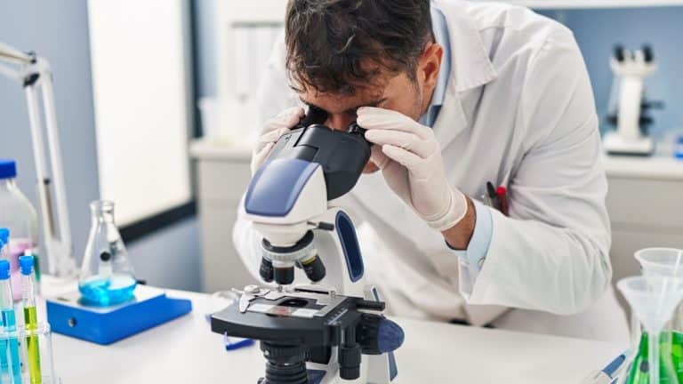 Mężczyzna w laboratorium patrzy przez mikroskop, otoczony prob&oacute;wkami i sprzętem laboratoryjnym.