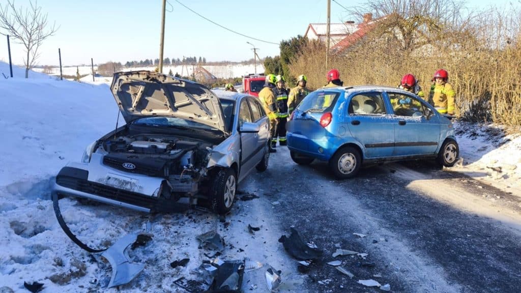 Zdjęcie z miejsca wypadku, dwa uszkodzone samochody na śliskiej drodze, strażacy w kaskach i odzieży ratunkowej.