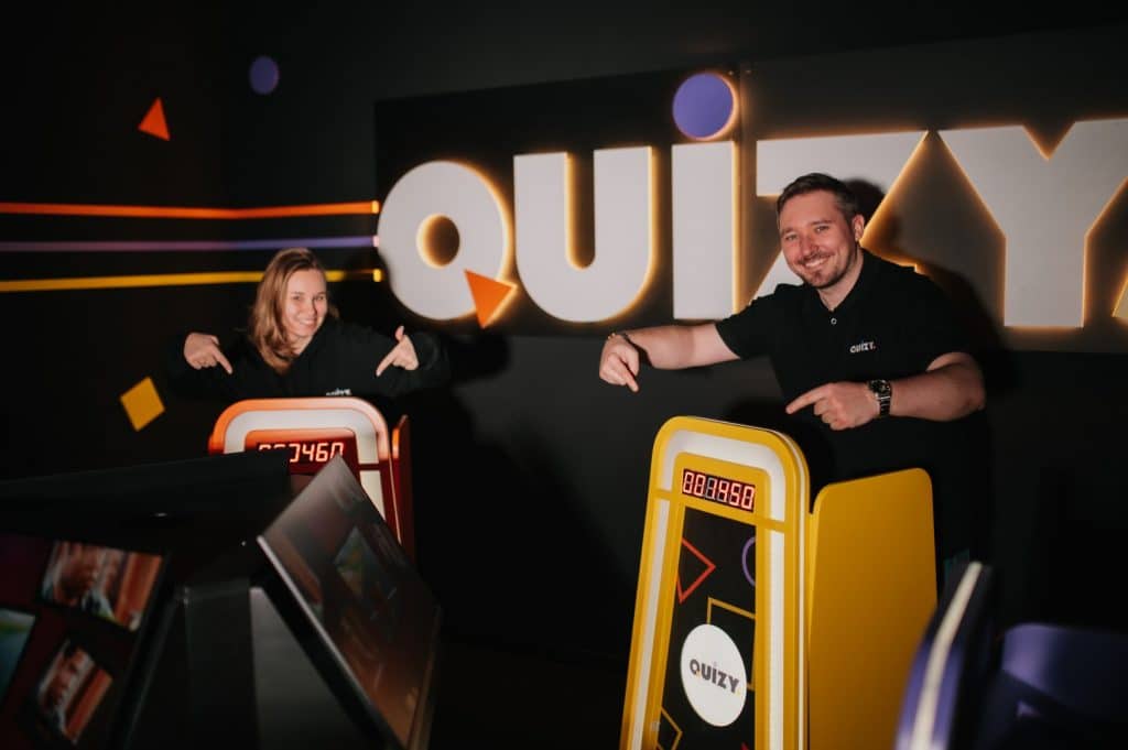 Dwoje uśmiechniętych ludzi wskazuje na plansze do quizu w nowym centrum rozrywki w Olsztynie.