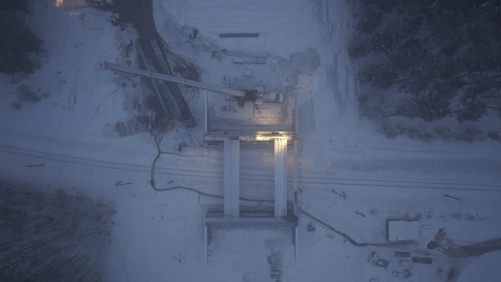 Widok z powietrza na plac budowy w zimowej scenerii, z widocznym ciężkim sprzętem i konstrukcjami.