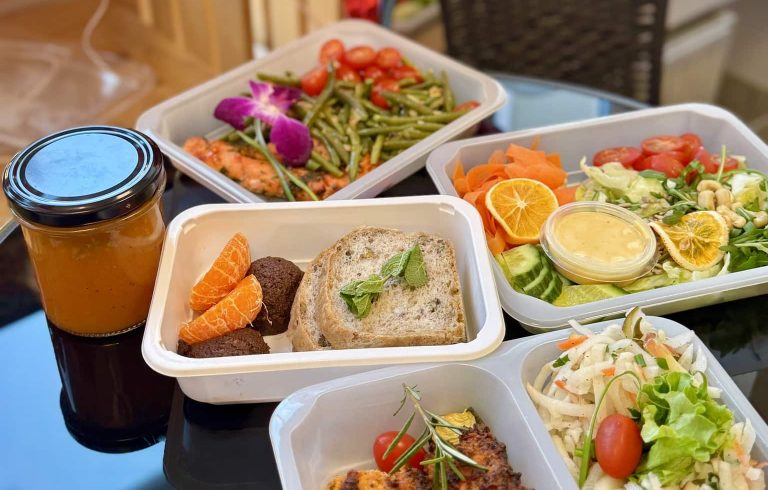 Zestaw zdrowych posiłk&oacute;w w pojemnikach, w tym sałatki, pieczywo i napoje, na tle ciemnego blatu.