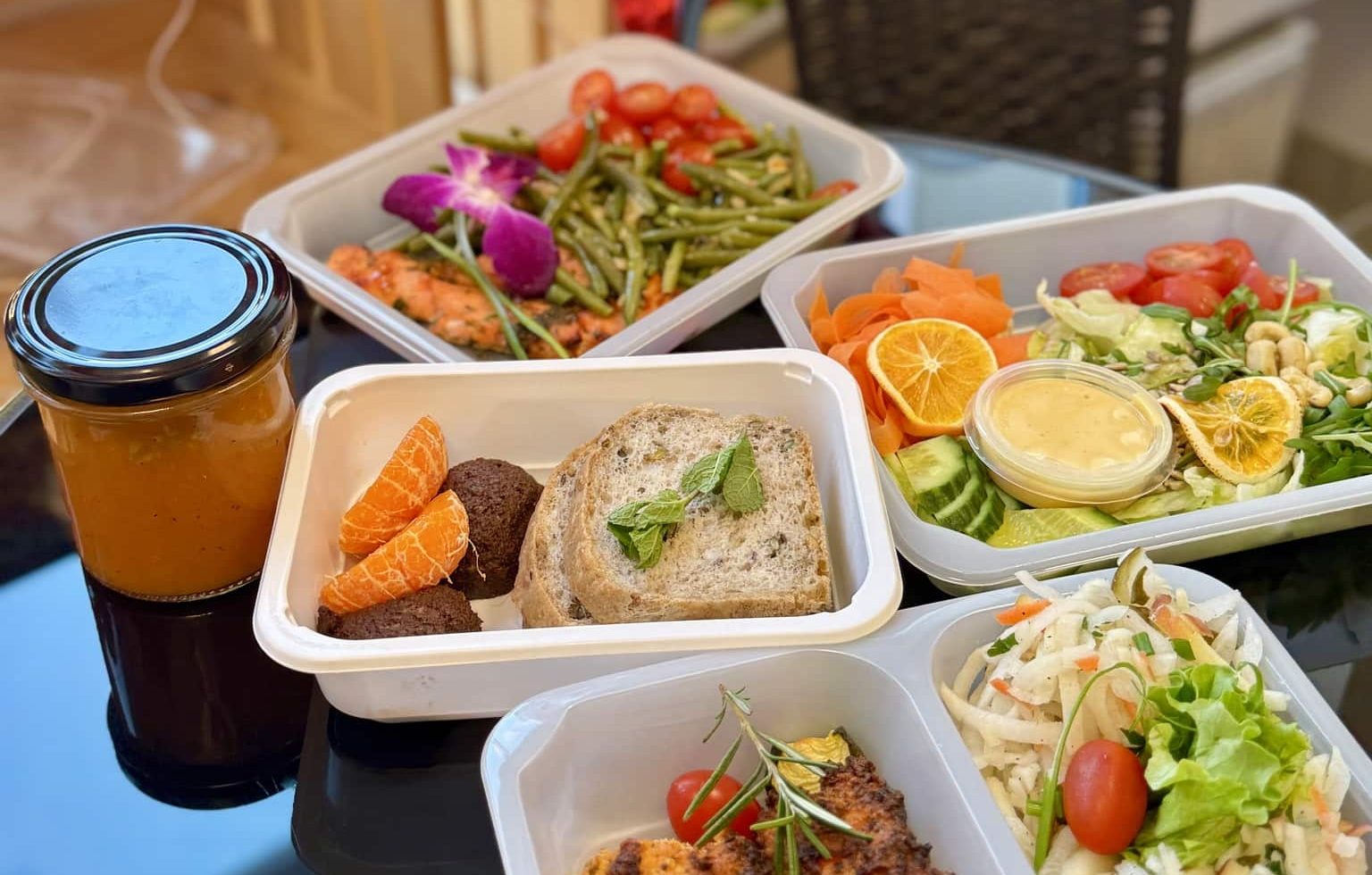 Zestaw zdrowych posiłk&oacute;w w pojemnikach, w tym sałatki, pieczywo i napoje, na tle ciemnego blatu.