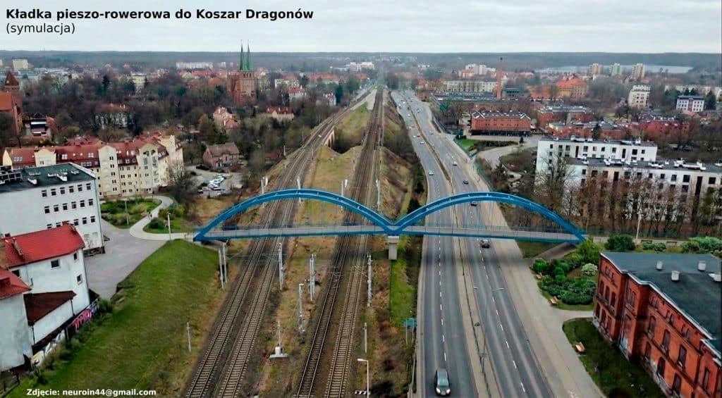 Widok na miasto z mostem nad torami kolejowymi i ulicami, w tle zabudowa i lasy.