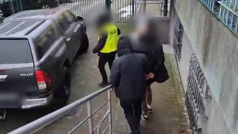 Grupa os&oacute;b w policyjnym pojeździe, w tle samoch&oacute;d, na mokrej ulicy, w trakcie działań służb.