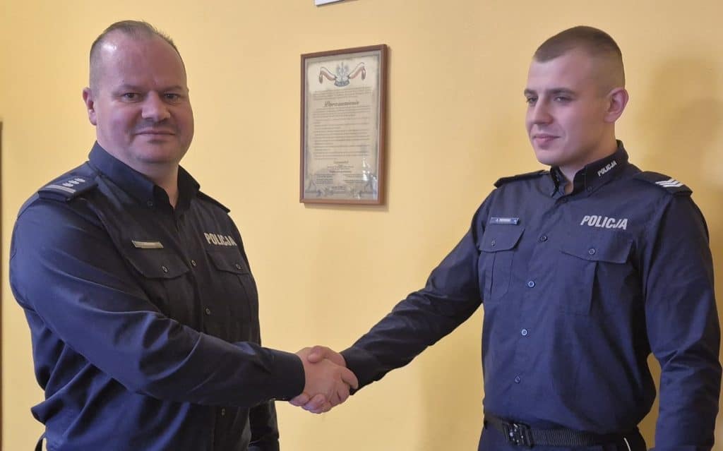 Policjanci podczas oficjalnej uroczystości, uściski dłoni, w tle ż&oacute;łta ściana i dokument w ramce.